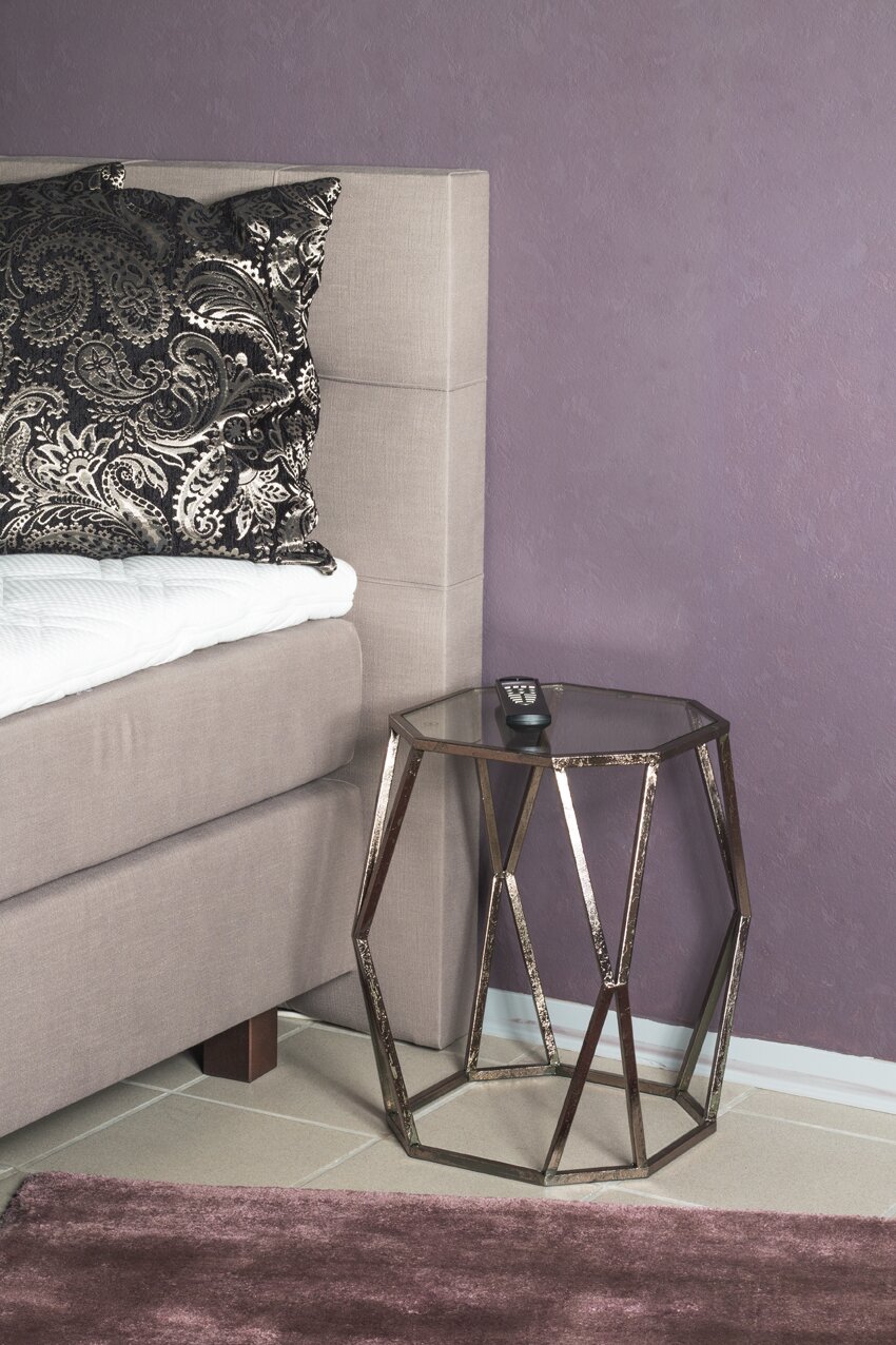 Moderner, geometrischer Beistelltisch aus Metall mit Glasplatte neben einem gepolsterten Bett vor lila Wand, seitliche Perspektive auf das Produkt.