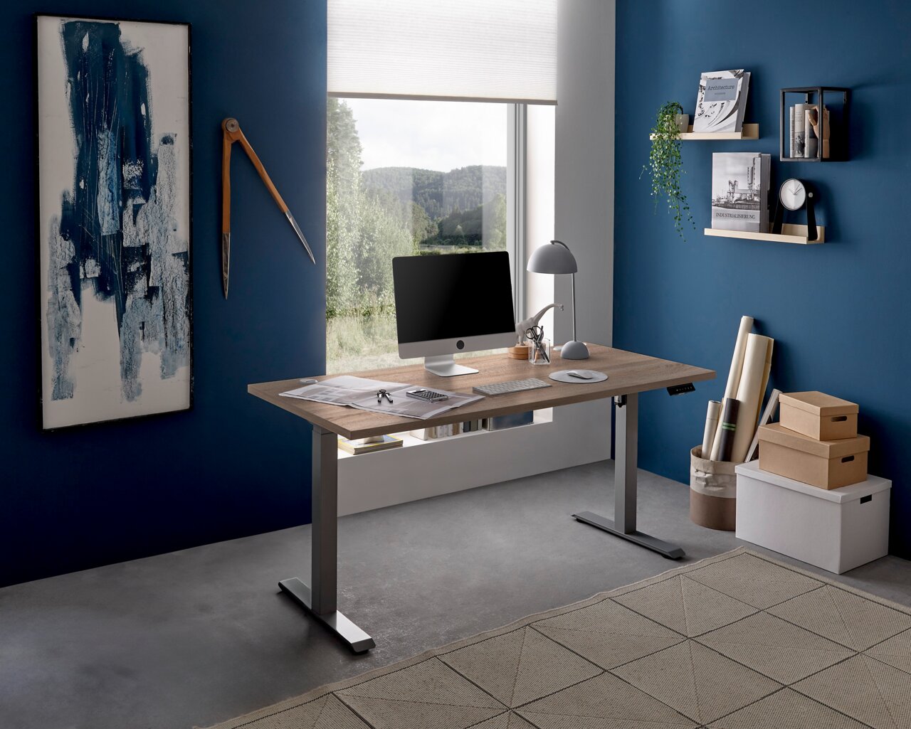 XORA E-bureau FINITO Modern elektrisch bureau in een stijlvol kantoor met een blauwe muur, gezien vanuit de zijkant. Het bureau heeft een houten blad en zilveren poten, en is voorzien van een computer en een bureaulamp.