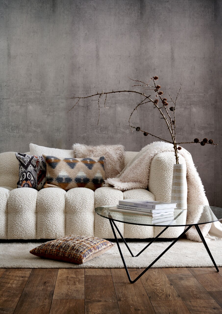 Wohnzimmer mit gemütlichem Sofa, dekoriert mit verschiedenen Wendekissenhüllen in neutralen und gemusterten Designs, im Vordergrund ein moderner Glastisch. Perspektive: Frontalansicht.