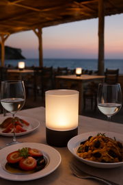 Akku-Windlicht auf einem Tisch bei Sonnenuntergang mit Meerblick, umgeben von Tellern mit Essen und Weingläsern, aus der Perspektive eines Gastes.