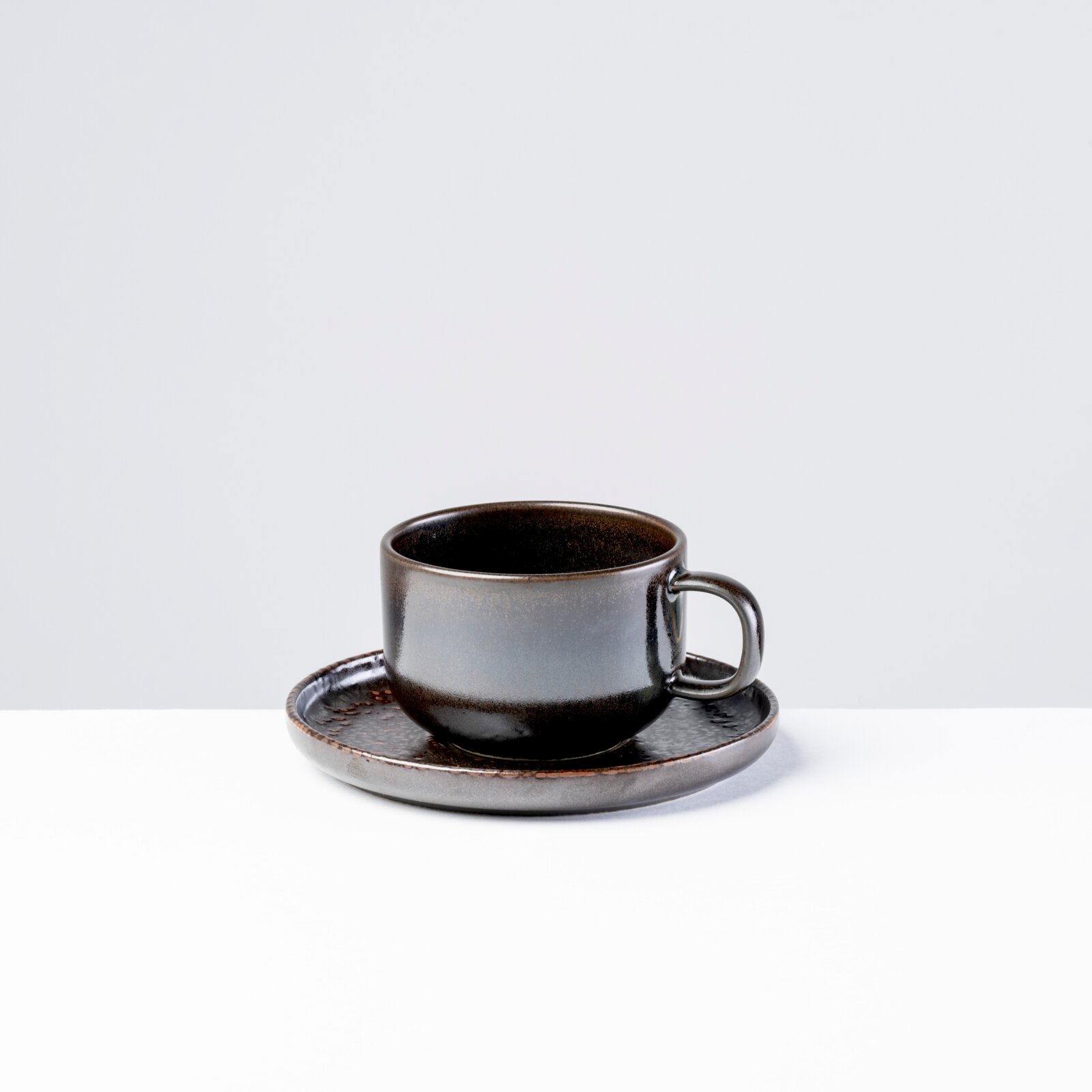 Dunkelgraue, glänzende Keramik-Kaffeetasse mit Henkel auf passender Untertasse vor hellem Hintergrund, frontal aus leicht erhöhter Perspektive