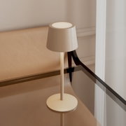 Beige, minimalistische led-tafellamp met een conische kap en een ronde voet op een glazen bijzettafeltje, gefotografeerd vanuit een licht bovenaanzicht/schuin aanzicht van voren