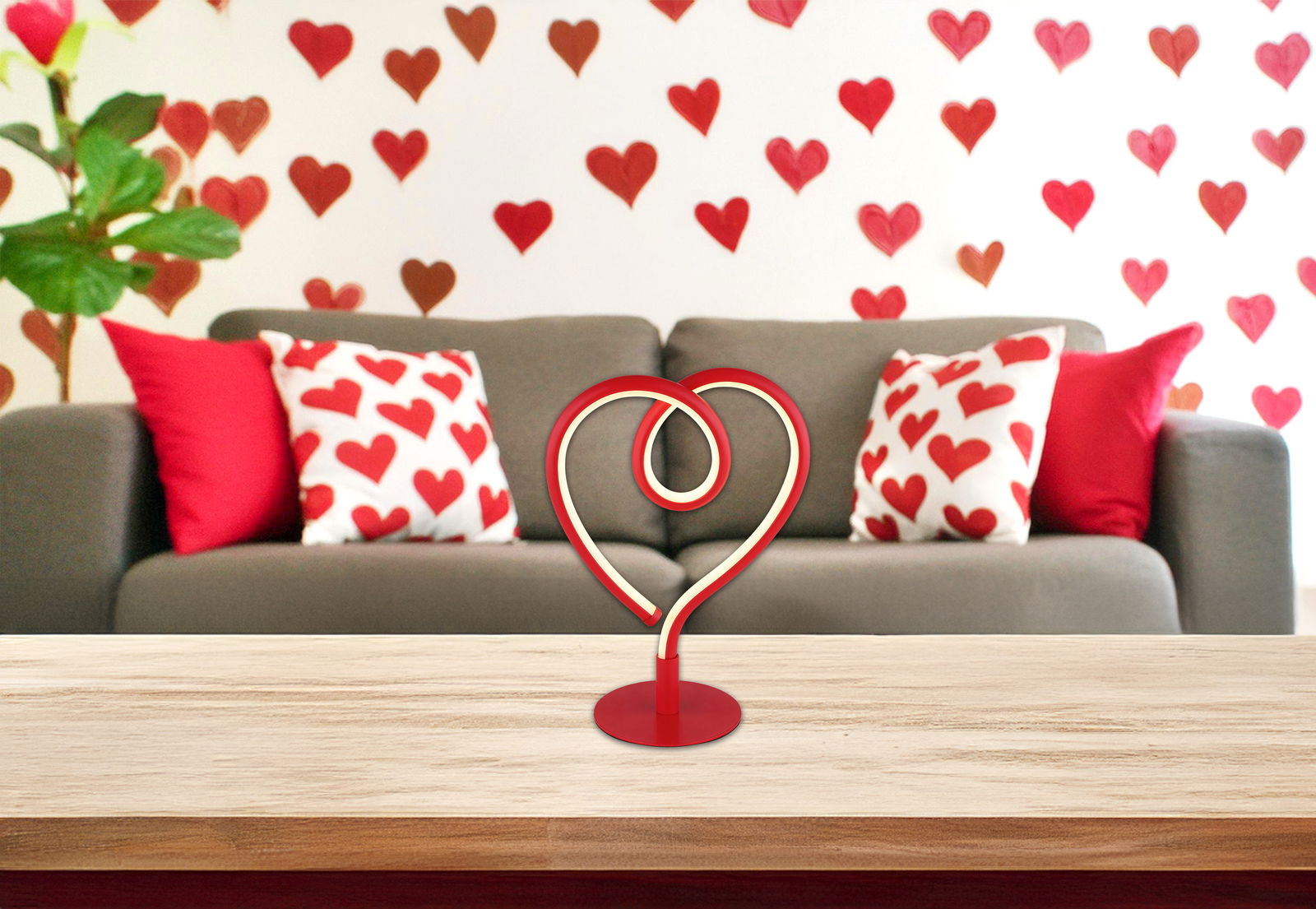 LED-Tischleuchte in Herzform auf einem Tisch, im Hintergrund ein Sofa mit herzgemusterten Kissen und herzförmiger Wanddekoration, frontal aufgenommen.