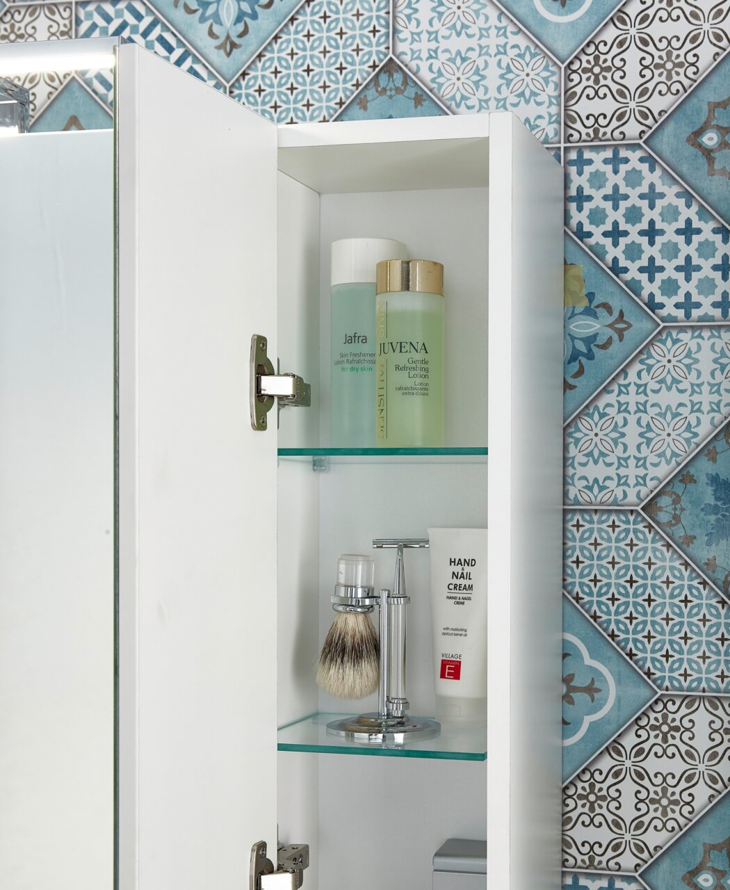 Geöffneter Spiegelschrank mit zwei Glasböden, gefüllt mit Pflegeprodukten und Rasierzubehör, vor einer dekorativen blauen Fliesenwand. Perspektive von vorne.