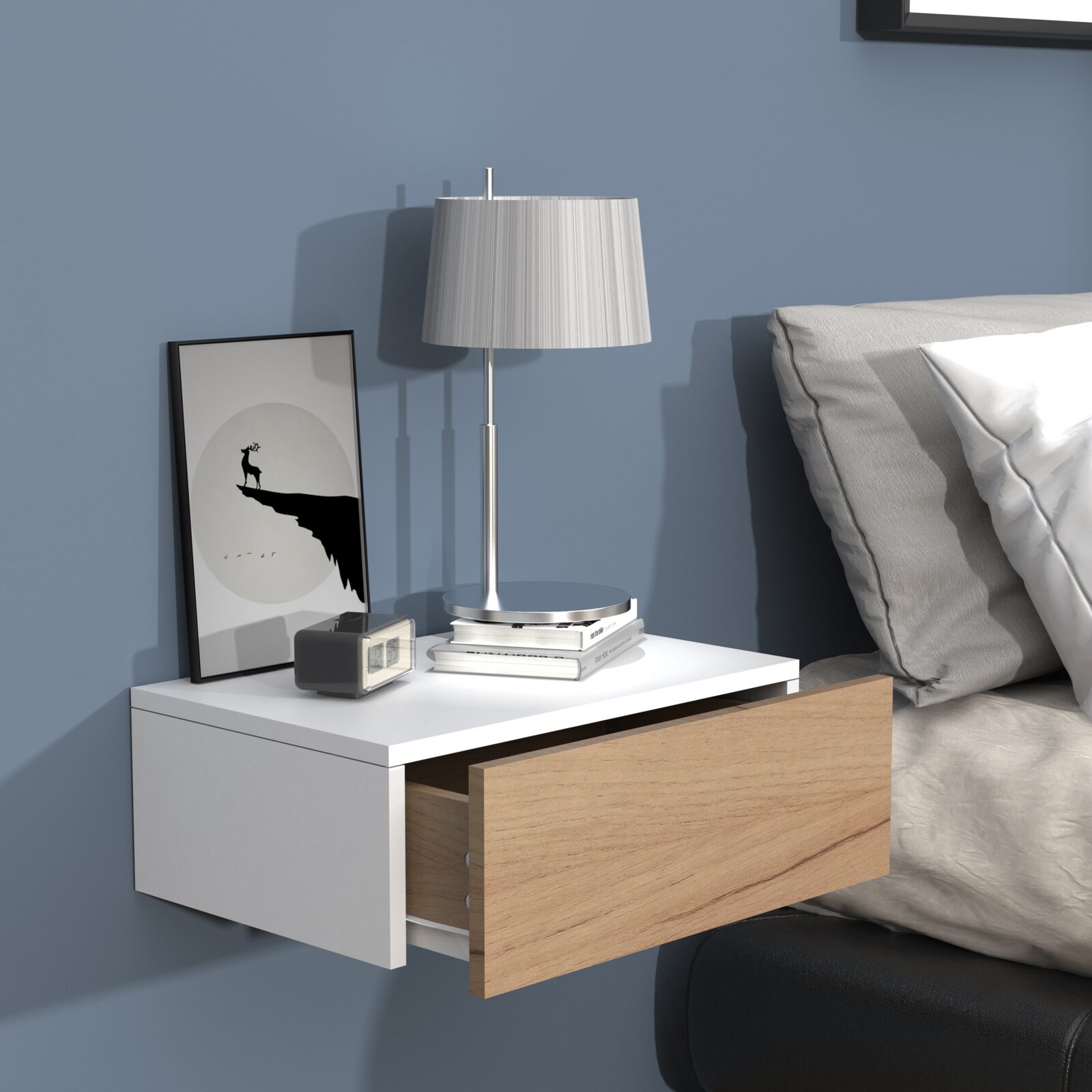 Wandmontierter Nachttisch in Weiß mit Holzschublade (halb geöffnet), darauf silberne Tischlampe und Bücher neben einem Bett – Perspektive: leicht schräg von vorne.