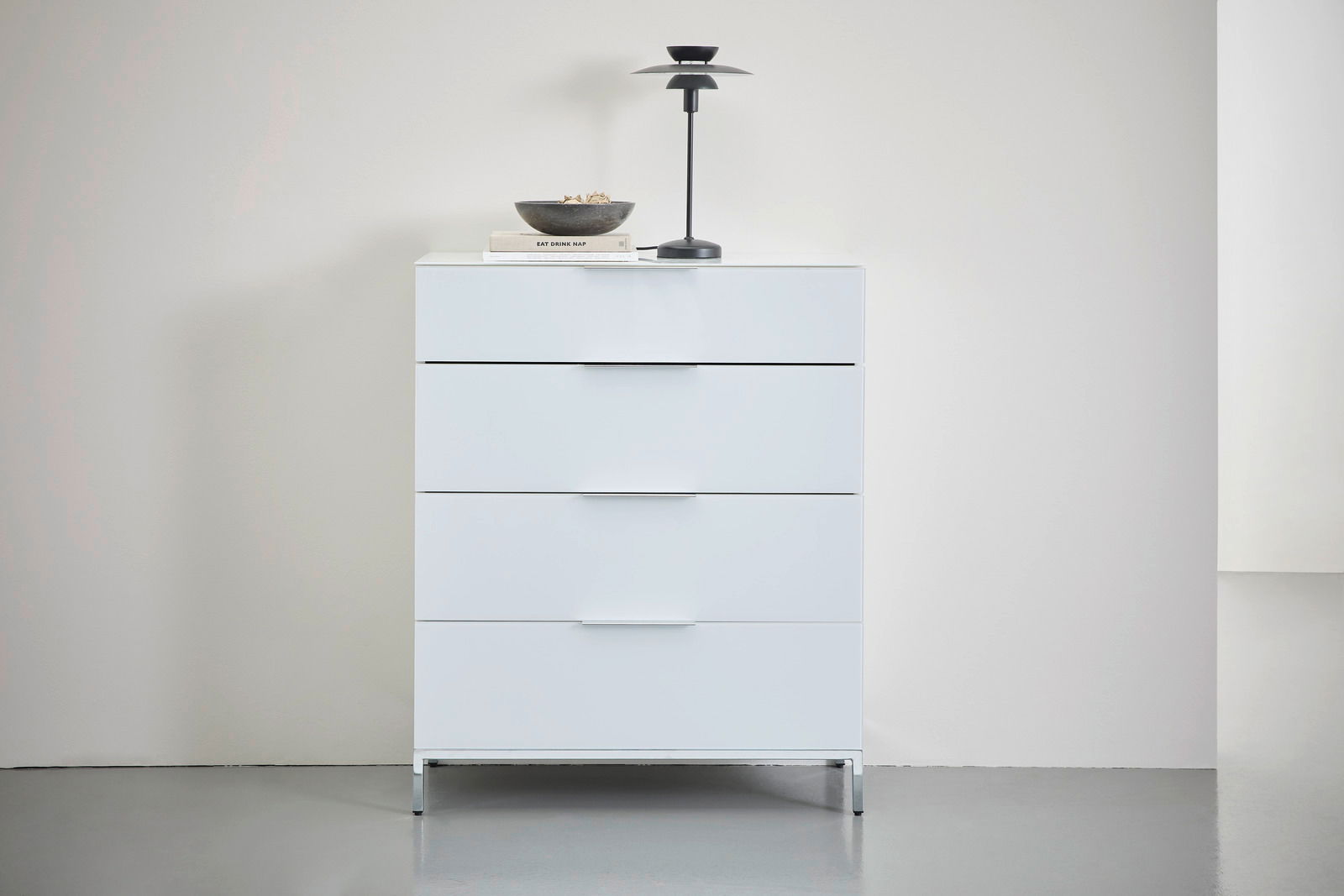 Weiße Schubkastenkommode mit vier Schubladen, dekoriert mit einer Lampe und einer Schale, aus frontaler Perspektive fotografiert.