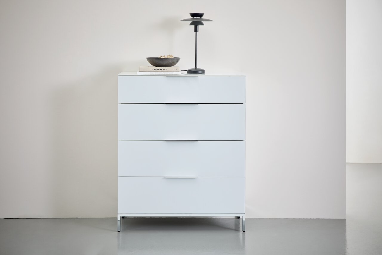 Weiße Schubkastenkommode mit vier Schubladen, dekoriert mit einer Lampe und einer Schale, aus frontaler Perspektive fotografiert.