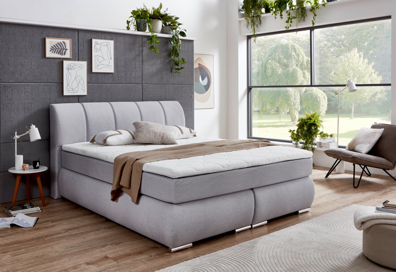 Intro Boxspringbett Modernes Boxspringbett in einem stilvollen Schlafzimmer mit grauem Kopfteil, dekorativen Kissen und Tagesdecke, seitliche Perspektive.