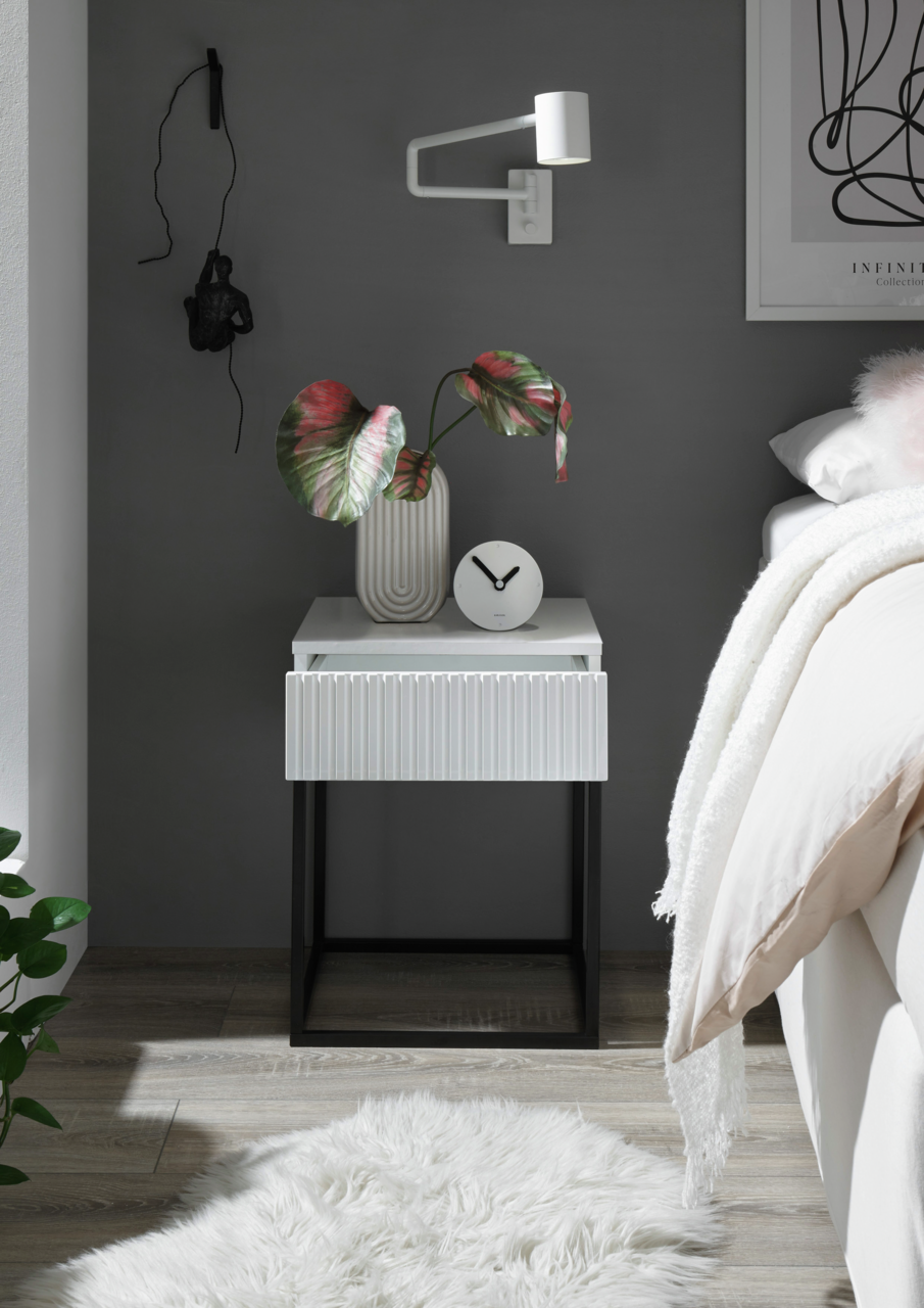 Moderne Nachtkommode mit weißer Oberfläche und geriffelter Schublade, dekoriert mit einer Vase und einer Uhr, aus frontaler Perspektive.