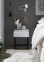Moderne Nachtkommode mit weißer Oberfläche und geriffelter Schublade, dekoriert mit einer Vase und einer Uhr, aus frontaler Perspektive.