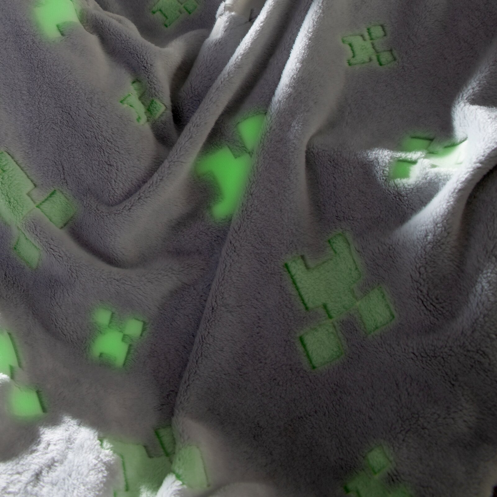 Nahaufnahme aus schräger Draufsicht auf eine weiche, graue Plüschdecke mit leuchtend grünen Pixel-/Creeper-Motiven, in Falten gelegt.