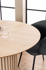 Runder Esstisch aus hellem Holz mit vertikalen Streben, fotografiert aus einer schrägen Vogelperspektive, mit zwei Gläsern und einem schwarzen Stuhl im Hintergrund.