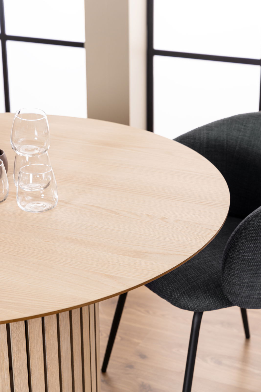 Runder Esstisch aus hellem Holz mit vertikalen Streben, fotografiert aus einer schrägen Vogelperspektive, mit zwei Gläsern und einem schwarzen Stuhl im Hintergrund.