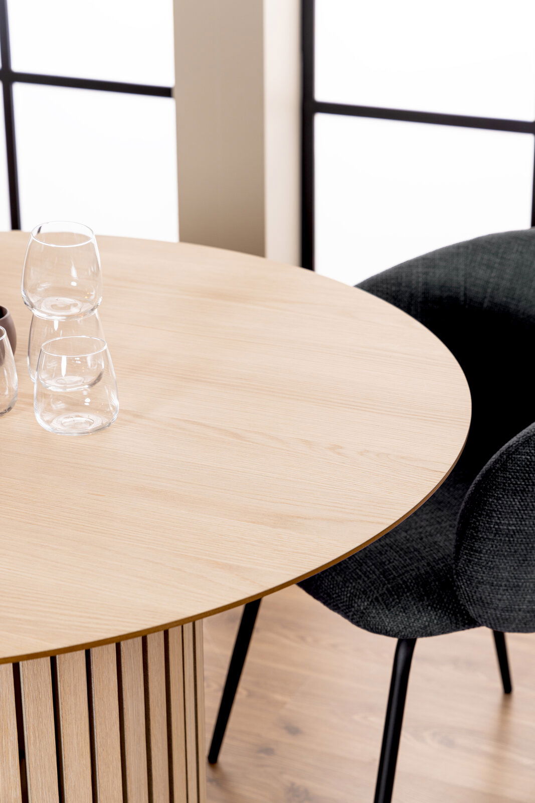 Runder Esstisch aus hellem Holz mit vertikalen Streben, fotografiert aus einer schrägen Vogelperspektive, mit zwei Gläsern und einem schwarzen Stuhl im Hintergrund.