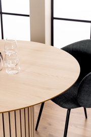 Runder Esstisch aus hellem Holz mit vertikalen Streben, fotografiert aus einer schrägen Vogelperspektive, mit zwei Gläsern und einem schwarzen Stuhl im Hintergrund.