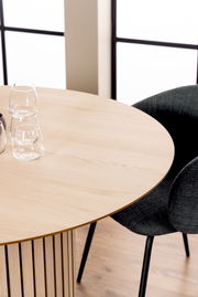 Runder Esstisch aus hellem Holz mit vertikalen Streben, fotografiert aus einer schrägen Vogelperspektive, mit zwei Gläsern und einem schwarzen Stuhl im Hintergrund.