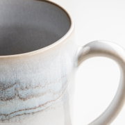 Detailaufnahme einer glasierten Keramik-Kaffeetasse in Weiß mit blau-brauner Sprenkel-Optik und Henkel, Perspektive: Nahaufnahme schräg von oben vor weißem Hintergrund