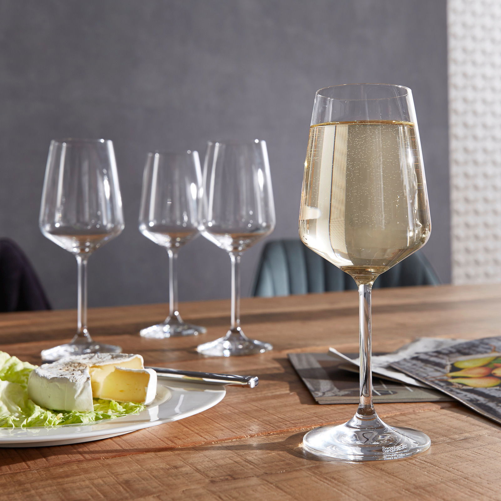 Spiegelau Weißweinglas Style 4er-Set Ein Set aus vier stilvollen Weißweingläsern auf einem Holztisch, eines davon gefüllt mit Weißwein im Vordergrund, mit einem Teller Käse und einem Messer daneben.