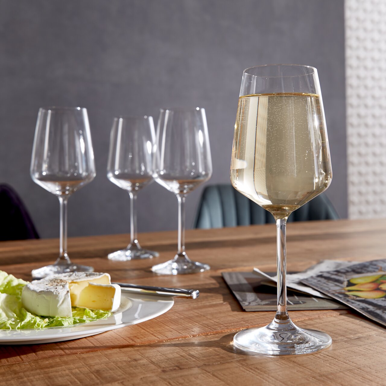 Spiegelau Weißweinglas Style 4er-Set Ein Set aus vier stilvollen Weißweingläsern auf einem Holztisch, eines davon gefüllt mit Weißwein im Vordergrund, mit einem Teller Käse und einem Messer daneben.
