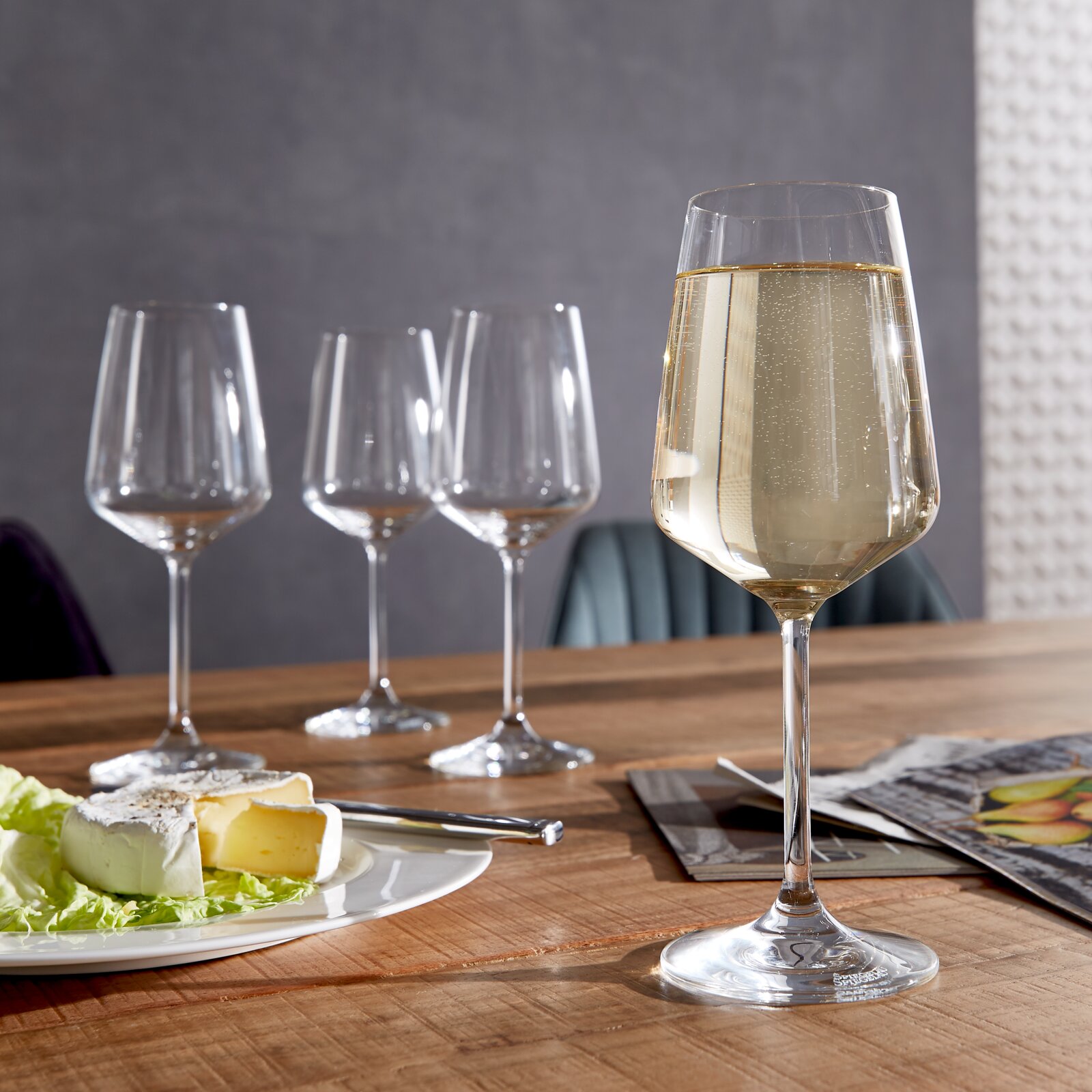 Ein Set aus vier stilvollen Weißweingläsern auf einem Holztisch, eines davon gefüllt mit Weißwein im Vordergrund, mit einem Teller Käse und einem Messer daneben.