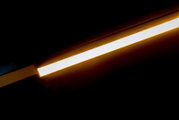 Schuine close-up zijaanzicht van een slanke, langwerpige LED-lichtbalk/plafondlamp met warmgele gloed tegen een donkere achtergrond