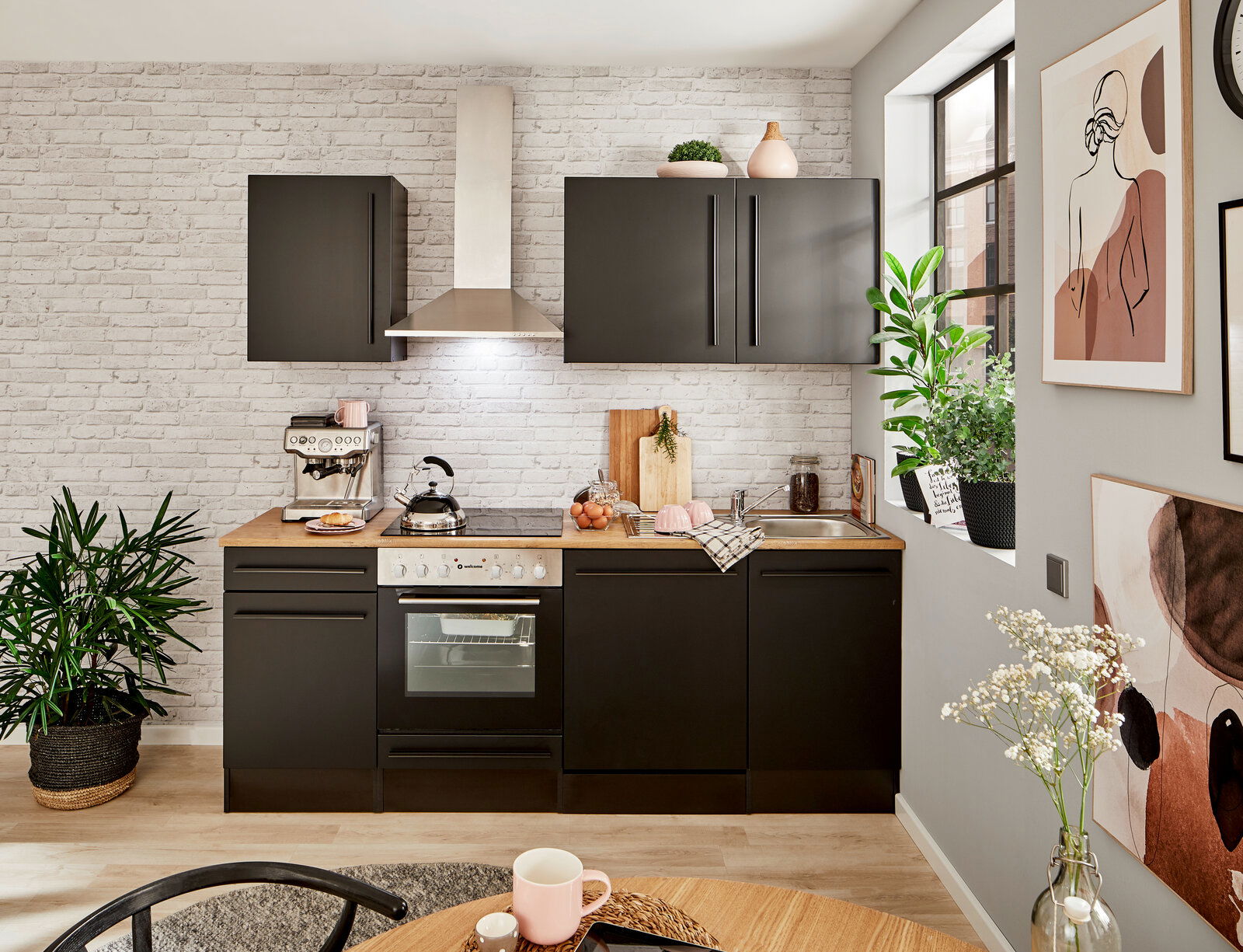 Moderne Küchenzeile in schwarzem Design mit Holzarbeitsplatte, Backofen, Herd und Dunstabzugshaube, umgeben von Pflanzen und Dekorationen, aus frontaler Perspektive.