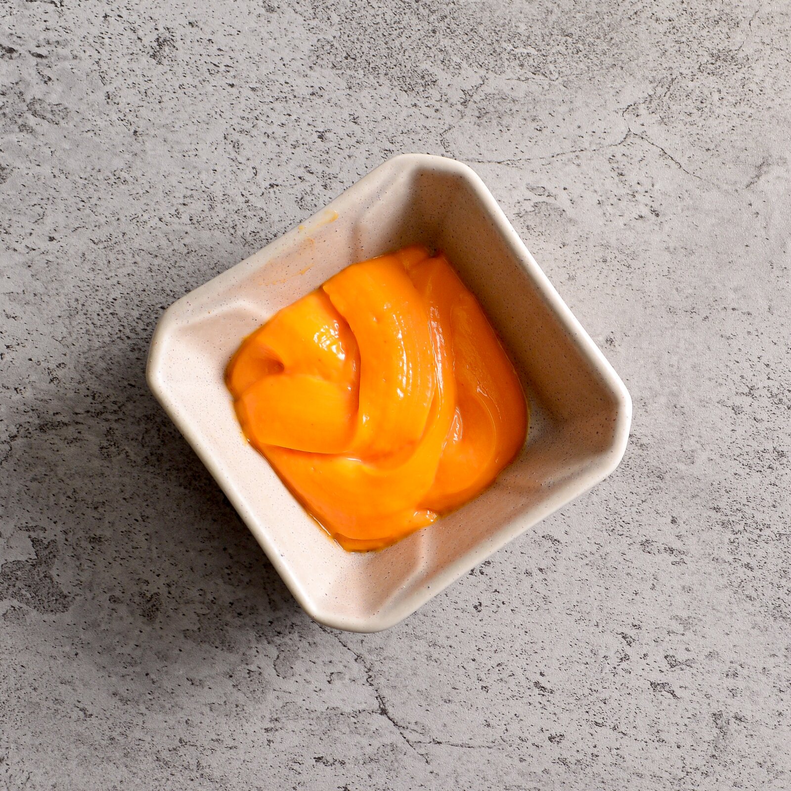 Quadratische, cremefarbene Keramikschale mit glänzendem orangefarbenem Püree auf grauer Steinoberfläche, aufgenommen aus der Vogelperspektive (Top-Down).