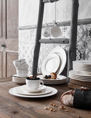 Ein stilvoll arrangiertes Geschirrset in Weiß, bestehend aus Tellern und Tassen, auf einem rustikalen Holztisch. Im Vordergrund eine Tasse mit Beeren, im Hintergrund gestapelte Teller und hängende Tassen. Seitliche Perspektive.