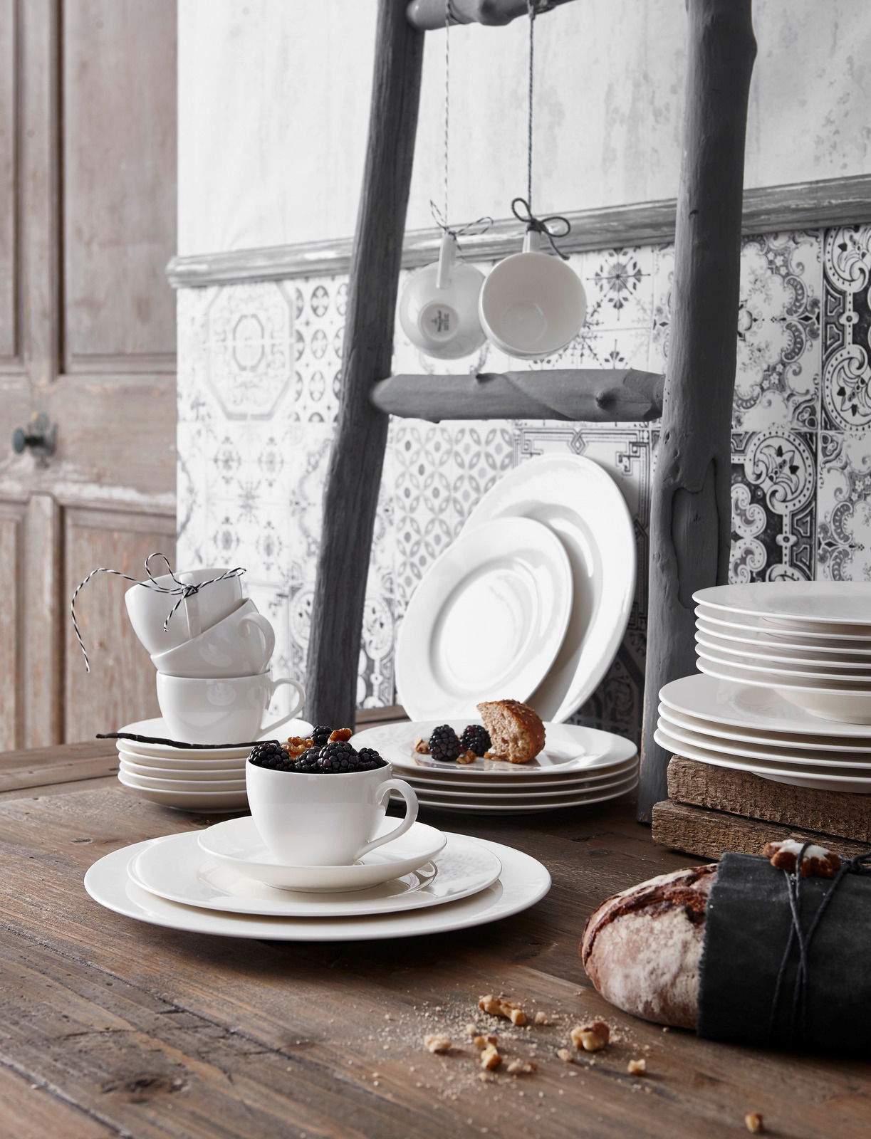 Ein stilvoll arrangiertes Geschirrset in Weiß, bestehend aus Tellern und Tassen, auf einem rustikalen Holztisch. Im Vordergrund eine Tasse mit Beeren, im Hintergrund gestapelte Teller und hängende Tassen. Seitliche Perspektive.