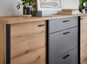 Nahaufnahme in leichter Schrägansicht von vorn: moderne Kommode/Sideboard aus hellem Holz mit grauen Schubladenfronten und schwarzen Griffen, dekoriert mit Pflanze, Büchern und Bildrahmen