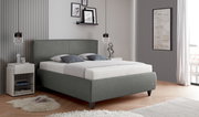 Modernes, grau gepolstertes Doppelbett mit hohem Kopfteil und schwarzen Füßen in einem stilvollen Schlafzimmer, schräg von vorn aus leicht seitlicher Perspektive aufgenommen.