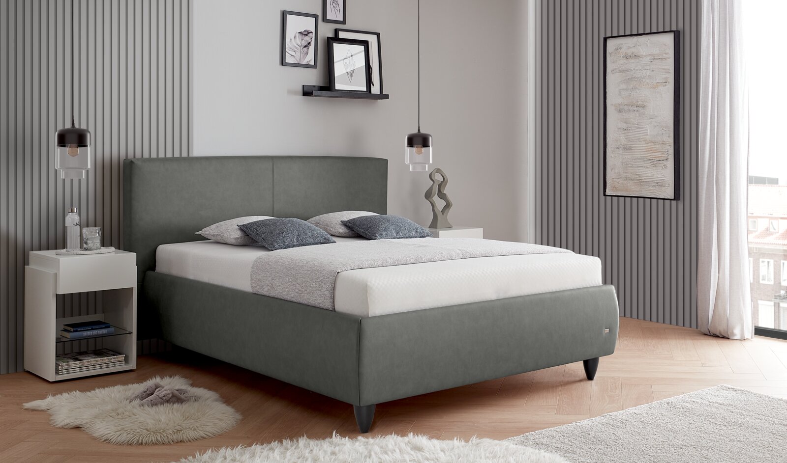 Modernes, grau gepolstertes Doppelbett mit hohem Kopfteil und schwarzen Füßen in einem stilvollen Schlafzimmer, schräg von vorn aus leicht seitlicher Perspektive aufgenommen.