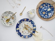 Flatlay aus Vogelperspektive: florales Geschirr-Set mit blau-weiß gemusterten Tellern, weißer Tasse und goldfarbenem Besteck auf hellem Hintergrund mit Holzbrett und Trockenblumen.