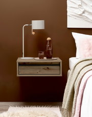 Schwebender Nachttisch aus hellem Holz mit Schublade neben einem Bett, darauf weiße Tischlampe und Glasflasche; frontale Ansicht auf Augenhöhe.