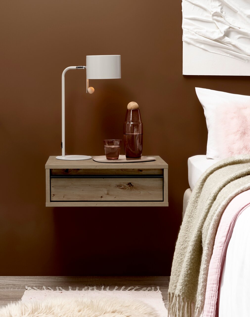 Schwebender Nachttisch aus hellem Holz mit Schublade neben einem Bett, darauf weiße Tischlampe und Glasflasche; frontale Ansicht auf Augenhöhe.