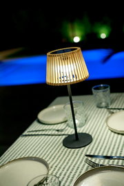 Leuchtende Tischlampe mit Holz-Lamellenschirm und schwarzem Standfuß auf gedecktem Tisch mit grün-weiß gestreifter Tischdecke, fotografiert aus leicht erhöhter schräger Perspektive.