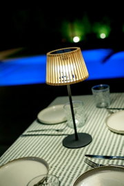 Leuchtende Tischlampe mit Holz-Lamellenschirm und schwarzem Standfuß auf gedecktem Tisch mit grün-weiß gestreifter Tischdecke, fotografiert aus leicht erhöhter schräger Perspektive.