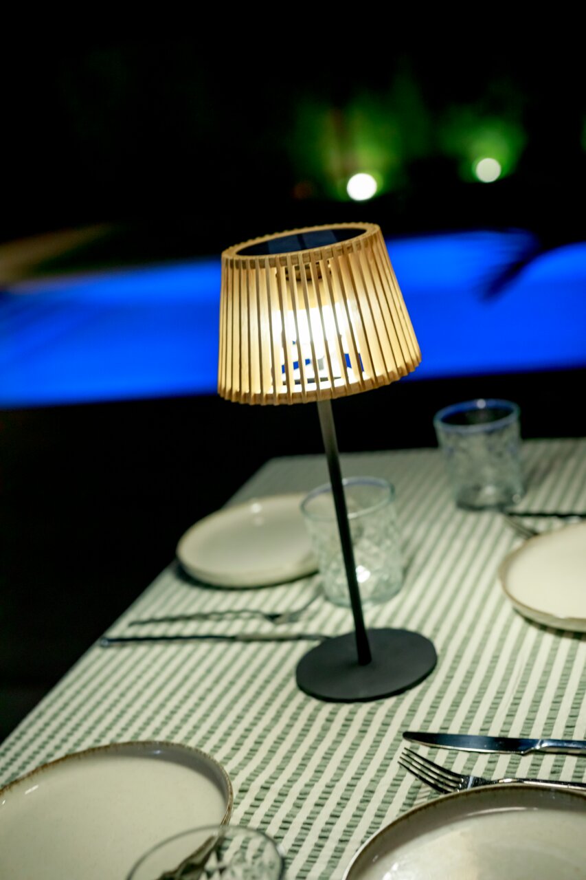 Leuchtende Tischlampe mit Holz-Lamellenschirm und schwarzem Standfuß auf gedecktem Tisch mit grün-weiß gestreifter Tischdecke, fotografiert aus leicht erhöhter schräger Perspektive.