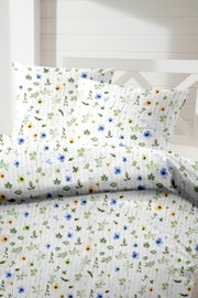 Blumige Bettwäsche in Weiß mit zartem botanischem Muster aus grünen Blättern sowie blauen und gelben Blüten, mit Kissenbezügen auf einem weißen Bett – Frontansicht leicht von oben