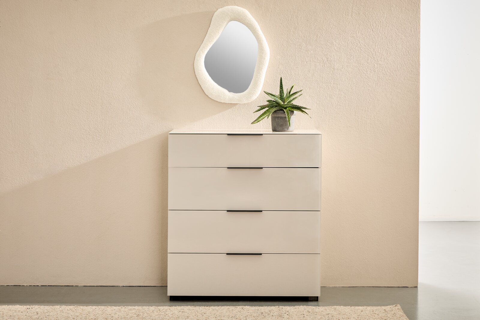 Novel Ladekast MALIN 2.0 Moderne, crèmekleurige ladekast met vier lades en zwarte handgrepen tegen een beige muur, met daarop een groene potplant en daarboven een organisch gevormde wandspiegel; vooraanzicht.
