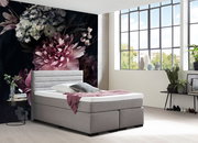 Graues Boxspringbett in modernem Schlafzimmer mit großen Fenstern und floralem Wandbild, seitliche Perspektive