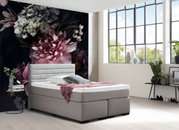 Graues Boxspringbett in modernem Schlafzimmer mit großen Fenstern und floralem Wandbild, seitliche Perspektive