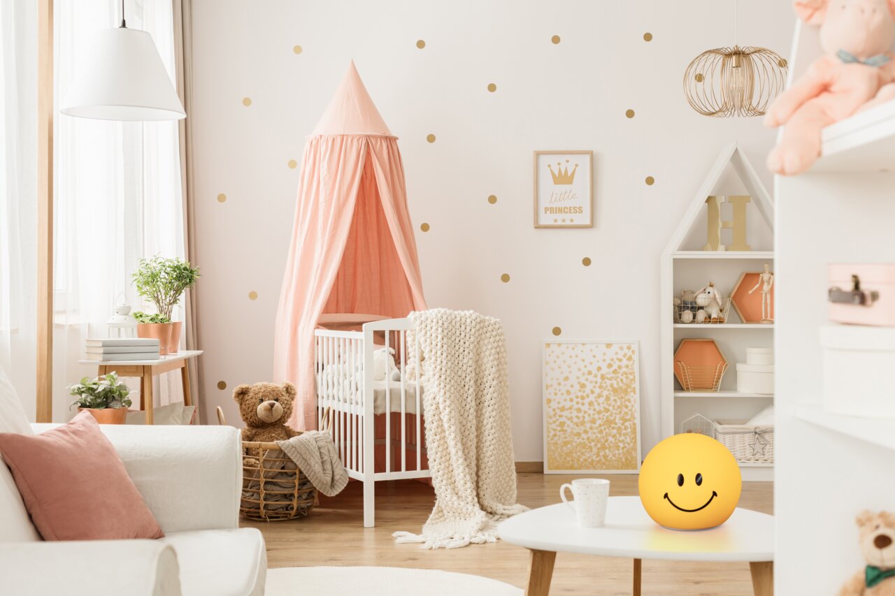 FHL easy Akku-Nachtlicht Smiley GLOWMOJI Akku-Nachtlicht in Form eines gelben Smiley-Gesichts auf einem Tisch in einem gemütlichen Kinderzimmer, seitliche Perspektive.