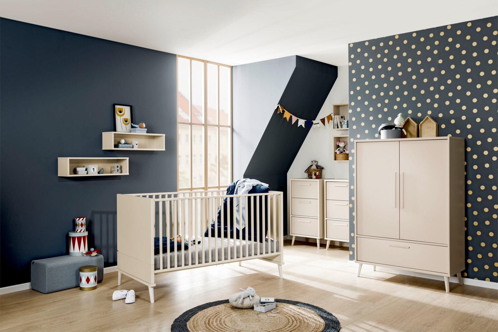 Modernes Babyzimmer mit cremefarbenem Kinderbett, Kommode und Kleiderschrank vor dunkelblauer Wand und gepunkteter Tapete, aufgenommen in einer weiten Raumansicht aus leicht erhöhter Perspektive schräg von vorne.
