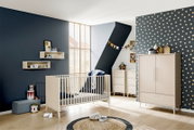 Modernes Babyzimmer mit cremefarbenem Kinderbett, Kommode und Kleiderschrank vor dunkelblauer Wand und gepunkteter Tapete, aufgenommen in einer weiten Raumansicht aus leicht erhöhter Perspektive schräg von vorne.