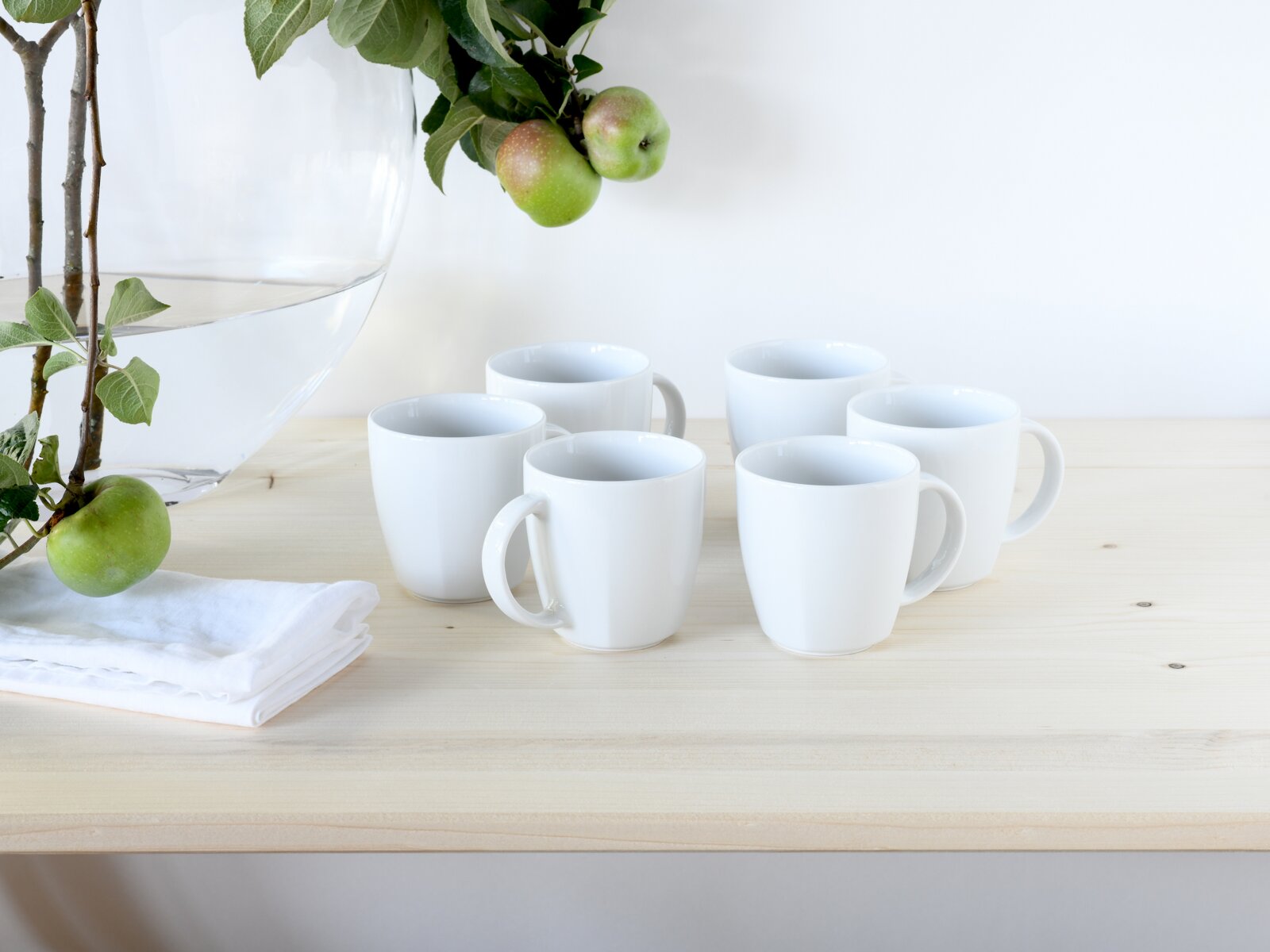 Fünf weiße Keramik-Kaffeetassen auf hellem Holztisch, links große Glasvase mit Apfelzweig und grünem Apfel; Perspektive: leicht erhöhte Frontansicht