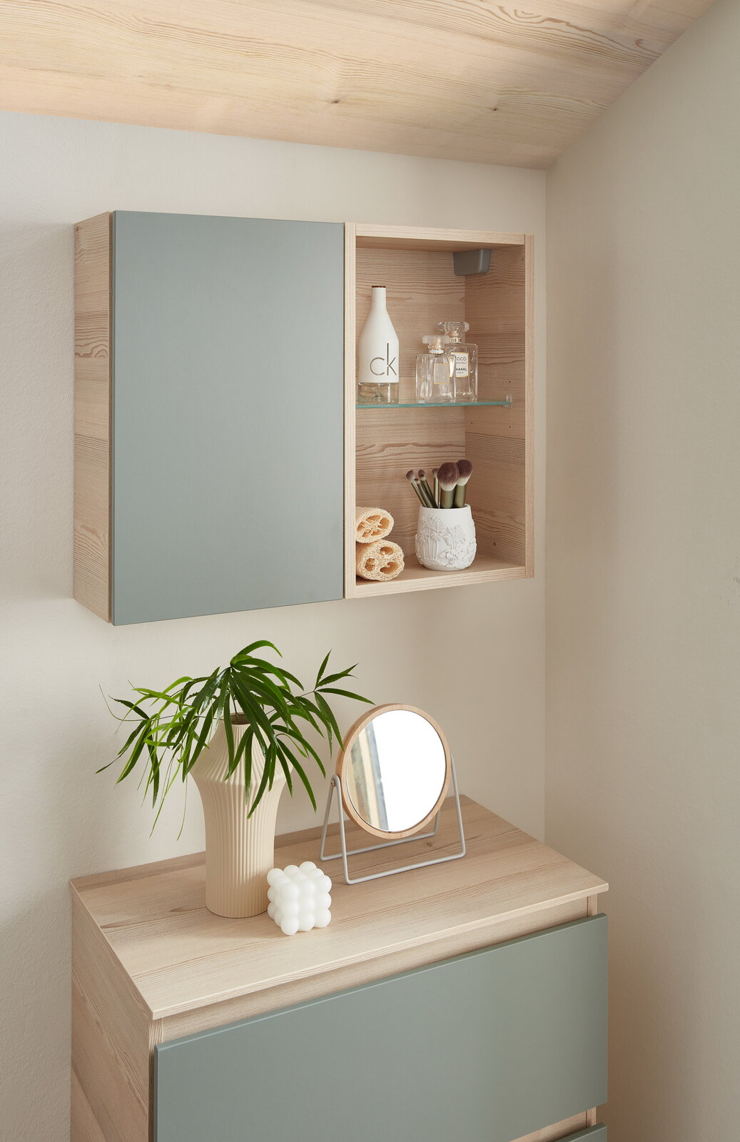 Hängeschrankregal in moderner Holzoptik mit einer geschlossenen und einer offenen Seite, gefüllt mit Dekorationsartikeln, aus einer seitlichen Perspektive fotografiert.