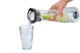 Hand gießt Wasser aus einer Wasserkaraffe BASIC 1L mit Limettenscheiben in ein Glas, seitliche Perspektive