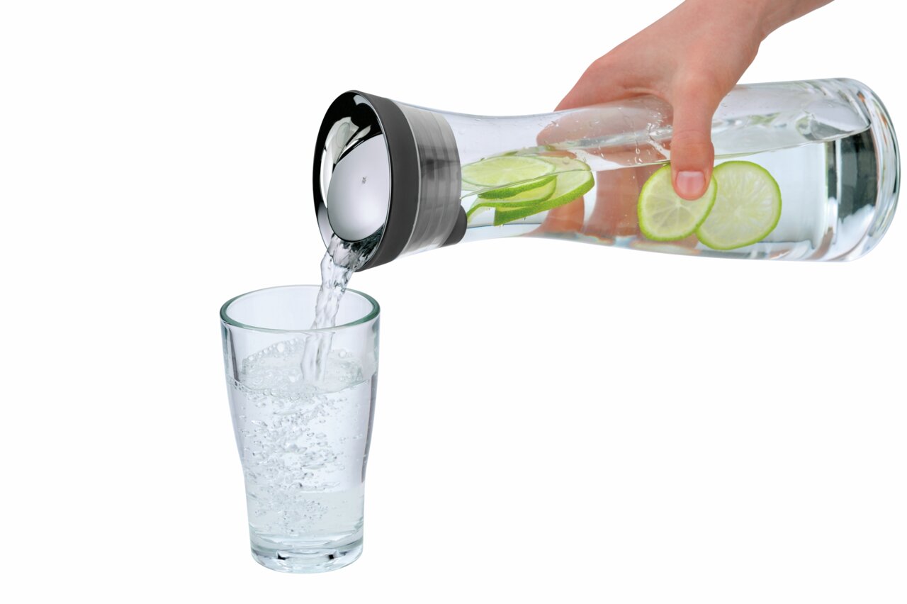 Hand gießt Wasser aus einer Wasserkaraffe BASIC 1L mit Limettenscheiben in ein Glas, seitliche Perspektive