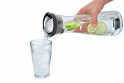 Hand gießt Wasser aus einer Wasserkaraffe BASIC 1L mit Limettenscheiben in ein Glas, seitliche Perspektive